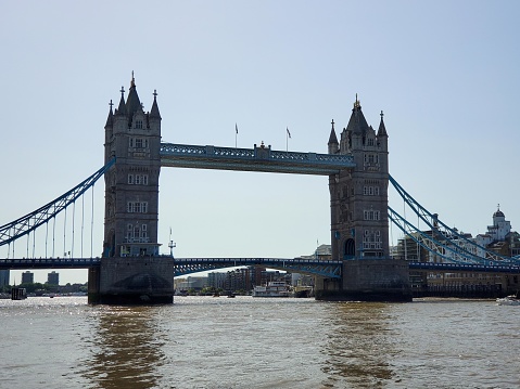 This is a photograph of the Tower Bridge in London, England. The bridge spans the River Thames with its two formidable towers and high-level walkways which are connected by two horizontal walkways at the upper level. It features Victorian Gothic style architecture with pointed towers capped with spires and flying buttresses. The bridge is painted in blue and white, and the bascules (the moveable sections of the bridge) appear to be down, allowing pedestrian and vehicular traffic to pass over. The sky is mostly clear with few clouds, suggesting a fair weather day, and the calm reflection of the bridge can be seen on the surface of the water.