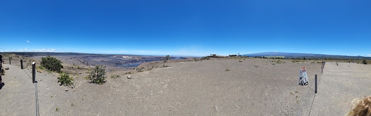 The picture provided is a panoramic view of a vast, open landscape under a clear, blue sky. The terrain appears to be a mix of barren and arid ground, with patches of low-lying shrubbery. In the middle distance, there is an elevated area with a structure that looks like an observation deck or visitor center, designed to blend with the natural surroundings. In the far background, a mountain or large hill looms, suggesting this location might be in a valley or near a volcanic area. There are protective fencing and signs likely indicating paths or important information for visitors. On the near right, out of focus, appears to be part of a person's head with blonde hair, suggesting the picture has been taken while in the presence of other people. The weather seems pleasant and suitable for outdoor activities.