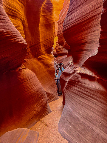 Page, Arizona, USA - September 21, 2023: Nestled within the desert of Northern Arizona, Antelope Canyon portrays the sheer beauty that nature can create. Famous for its lavish colors, unique patterns and complex textures, this slot canyon is a geological wonder that mesmerizes all who venture into its depths. Carved into the red sandstone of the Navajo Nation, the canyon comes alive with a vibrant spectrum of reds, oranges, and yellows. The sunlight filtering through the narrow openings above casts an ever-changing, warm glow on the smooth, curving walls, creating a surreal play of shifting light and dancing shadows. The rock formations within the canyon walls bear the unmistakable marks of time and weather. Millennia of wind and water erosion have sculpted the sandstone into flowing, sinuous shapes, creating a sensory experience that is as tactile as it is visual. Touching the canyon's surface connects a person directly to the forces of wind and water that have shaped it over countless millennia. In this photograph a couple is exploring the slot canyon. Antelope Canyon is in Coconino County near Page, Arizona, USA.