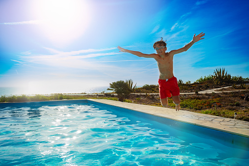 A boy in red swim shorts jubilantly leaps beside a sparkling pool on a sunny day