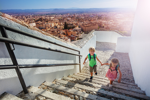 Two kids with backpacks climb up hill reaching viewpoint to admire Alhambra ancient palace on their summer trip to Granada, Spain