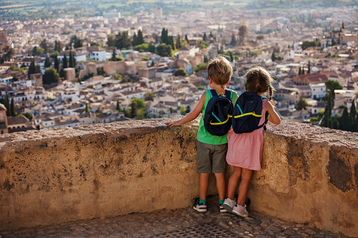 Brother and sister with backpacks standing on viewpoint enjoy Alhambra ancient citadel view on their summer family trip to Granada, Spain