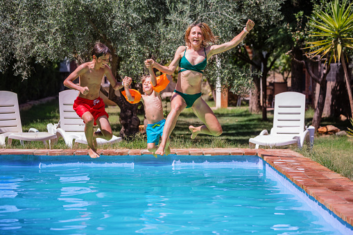 Happy mother wearing a green two-piece swimsuit and her to sons energetically jump into the pool, with one arm raised in a gesture of delight