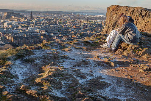 Edinburgh, Scotland - Jan 17, 2024 - Two young women sitting relaxing on top of rock over the mountain and enjoying looking at scenic view of cityscape. Two female Sitting down enjoying the amazing view from the mountain top of Salisbury Crags, Destinations in Europe, Space for text, Selective focus.
