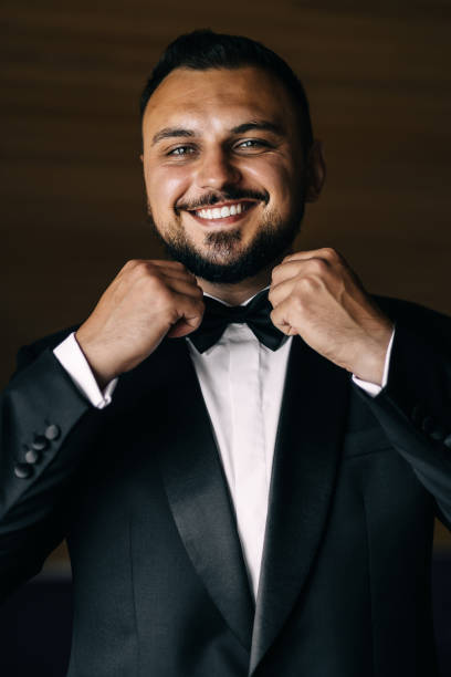 istock Jolly groom standing and fixing his black tie while looking and smiling to the camera. 1998903560