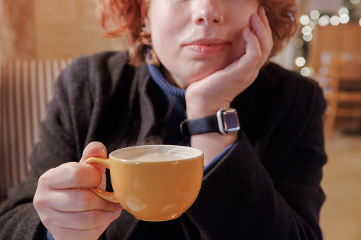 In a cafe, an unrecognizable redhead woman drinks hot coffee in a yellow cup