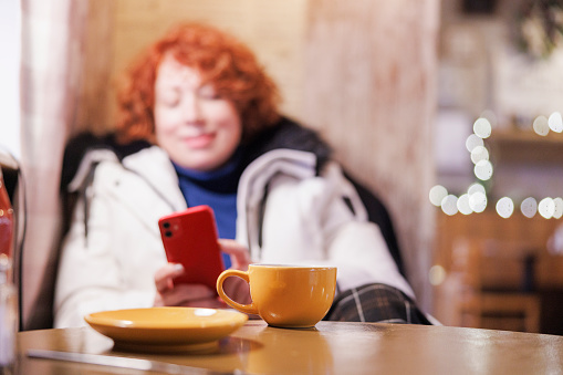 Empty yellow cup and plate after the meal in a restaurant. Woman with red hair hangs on her phone on background