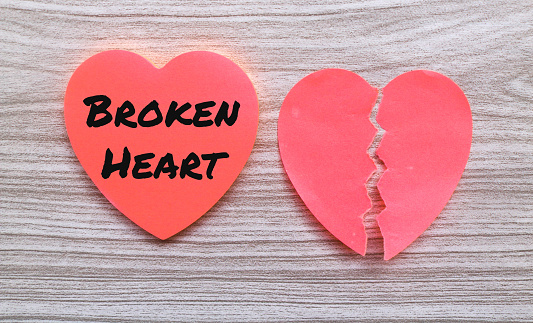 The words broken heart on a sticky note in the shape of a torn red heart isolated on a gray wooden background. Broken heart concept