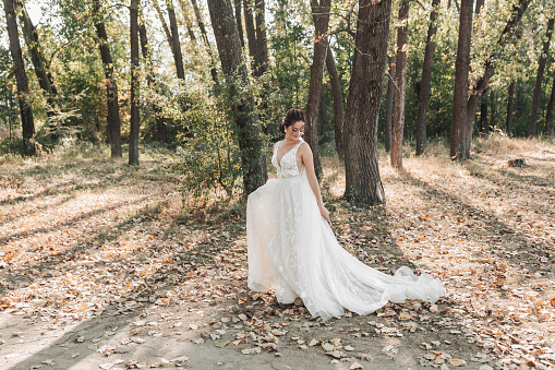 The bride in a white dress with a long train is walking in the forest, looking down. The wind blows the girl's hair. The shadows Summer wedding.