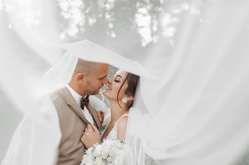 Wedding portrait of the bride and groom. Happy bride and groom gently hug each other under the veil, pose and kiss. Stylish groom. Beautiful young bride