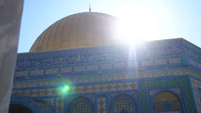 Camera pans from behind a pillar to reveal the Dome of the Rock as the sun shines behind the dome. You can see the beautiful architectural details on the building on the temple mount.