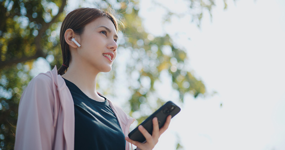 Low angle view, Selective focus, Young asian woman wear wireless earphones is resting listening the music from her smartphone holding and happily in public park, outdoor exercise concept, health care