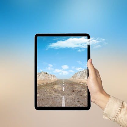 Human hand holding tablet with a screen view of a street with hills and rock cliff view with blue sky. Traveling concept