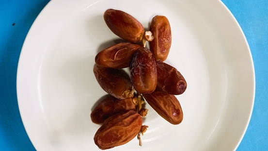 pile of dates with stems on a white plate, dates usually eaten to break the fast in the month of Ramadan, isolated on blue background