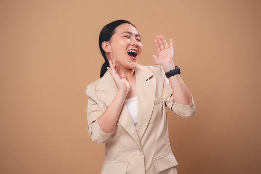Asian woman happy smiling announcement making a telling shouting gesture, sharing news, standing isolated on beige background.