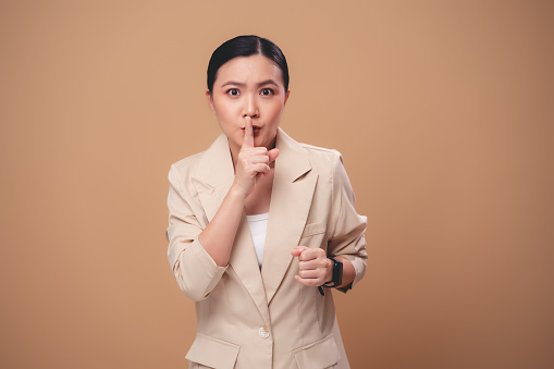 Asian woman annoyed frowning and putting index finger on lips meaning keeping silence, standing isolated on beige background.
