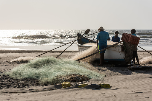 Morjim, Goa, India, 02.23.2017. Three fishermen near a boat preparing fishing nets on the ocean shore