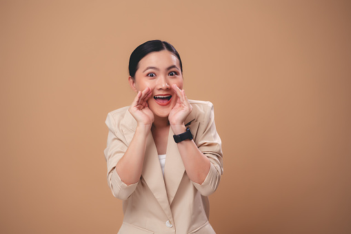 Asian woman happy smiling making a whispering gesture telling secret or sharing news standing isolated on beige background.