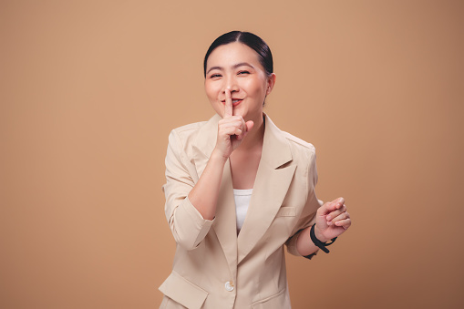 Asian woman happy smiling putting index finger on lips meaning keeping secret, standing isolated on beige background.