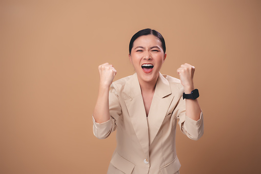 Asian woman happy smiling showing a winning gesture standing isolated on beige background.