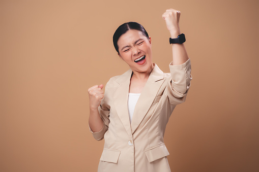 Asian woman happy smiling showing a winning gesture standing isolated on beige background.