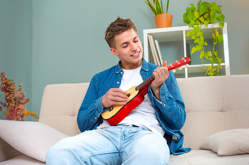 The young man plays the ukulele at home. Media. The man plays the ukulele alone at home. Young man practices playing a small guitar while sitting on the couch