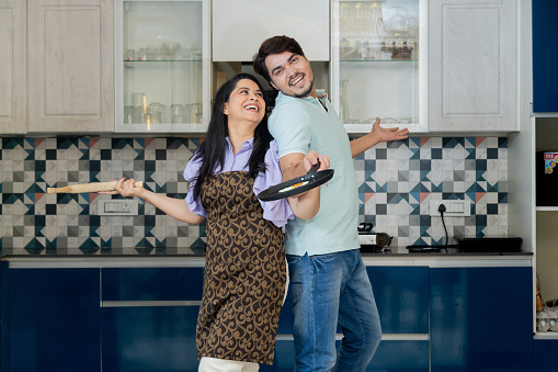 Happy indian couple having fun together in the kitchen, Wife wearing apron uniform with rolling pin in her hand and husband with non stick pan in his hand. love and relationship concept.