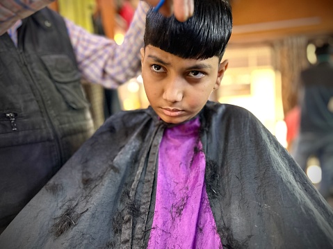 Indian little boy sitting on a chair and getting his hair cut by barber in the barber shop.