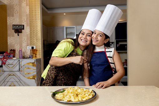 Happy indian mother and daughter dressed in cook uniform wearing a apron, chef hat ready to make tasty food with fresh vegetables. Standing in kitchen having fun.