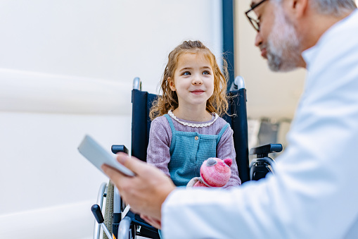 Friendly pediatrician talking to little patient in wheelchair. Cute preschool girl in wheelchair greeting doctor in hospital. Concept of children healthcare and emotional support for child patients.