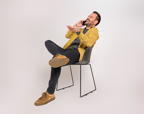 Cheerful young male entrepreneur relaxing on chair and talking over smart phone on white background