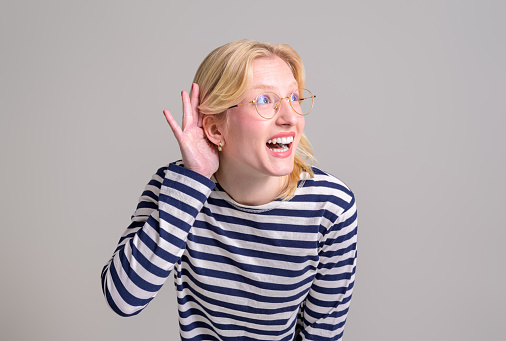 Curious happy blond young woman with hand near ear trying to listen gossip over white background