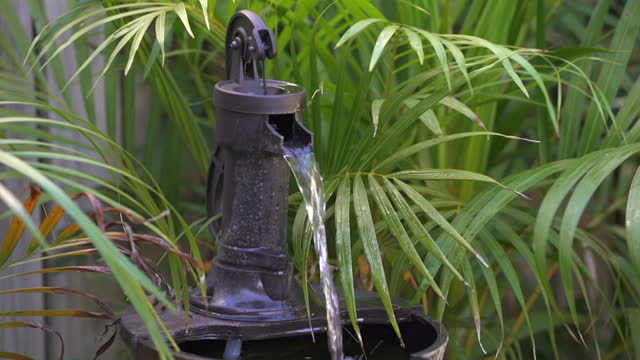 Cool fresh water flowing from an old water pump being used as a water feature fountain, surrounded by bright green palm leaves in a lush tropical garden.