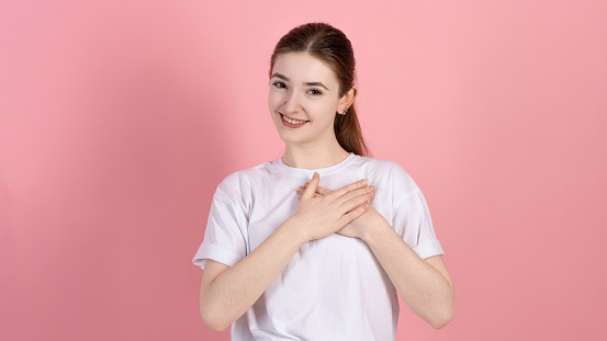 Portrait of a touched attractive Caucasian young brunette woman in a casual white t-shirt, touches the heart, the girl is grateful and says thank you, on a pink studio background