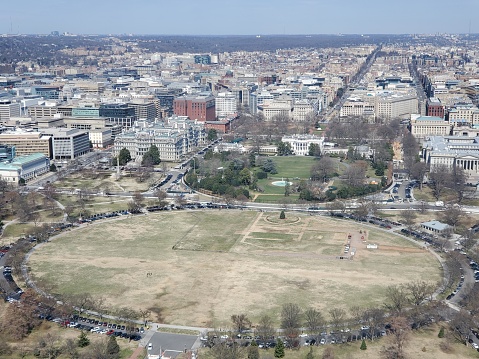 This picture appears to be an aerial view of a cityscape, likely taken from a high vantage point. In the foreground is a large, oval-shaped grassy area with paths that seem to divide it into quarters. There is a fountain in the center, and the oval is surrounded by a circular road with parked cars. Beyond the oval, the landscape is densely packed with buildings, roads, and patches of greenery. The architecture varies, with some buildings featuring red brick facades while others are more modern. The day is clear, and the view extends to the horizon where the sky meets the urban sprawl. This photograph might be of a significant location or park in a metropolitan area, possibly in Washington D.C., given the layout's resemblance to The Ellipse, a park south of the White House.