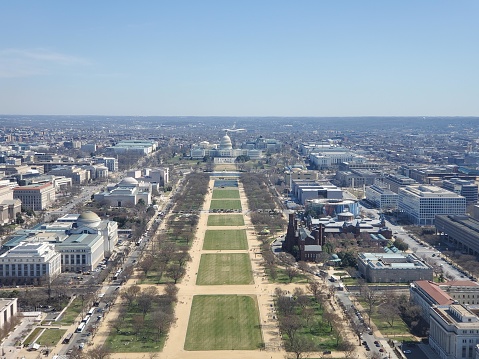 This is an aerial photograph showing a view over the National Mall in Washington, D.C., looking eastward. In the foreground, you can see wide, open grassy areas with walking paths. There are clusters of trees scattered throughout, and several large, stately buildings flanking either side of the Mall, which are some of the Smithsonian museums and federal institutions.   In the distance, you can see the United States Capitol building with its iconic dome. The sky is clear and bright, indicating it's likely a sunny day. The city stretches out all around the National Mall, with various buildings of different sizes and architectures. The overall feeling is one of openness and grand, organized layout, typical of the Mall's design as a place for public gathering, events, and as a vista for some of the country's most important civic landmarks.