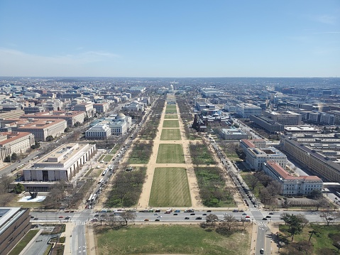 The picture shows an aerial view looking down a long, wide, grassy mall lined with trees and paths. This is the National Mall in Washington D.C., United States. On the left and right of the image, there are several large, white rectilinear buildings, which are likely some of the Smithsonian museums and government buildings. The photograph is taken on a clear day with a blue sky overhead, and you can see the cityscape extending into the distance with various buildings. The streets around the National Mall appear to be busy with vehicular traffic.