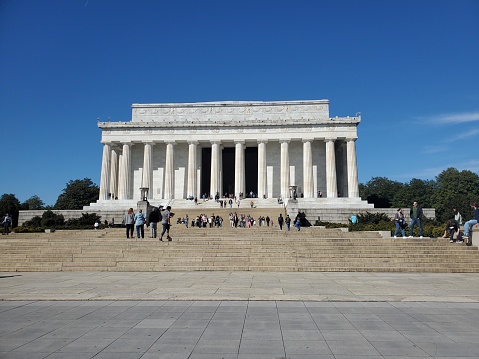 The picture shows the Lincoln Memorial, an iconic neoclassical monument located in Washington D.C., USA. It is a bright, clear day with blue skies. The memorial has a large number of white stone columns supporting the structure. In front of the memorial, there are steps leading up to the entrance where we can see visitors gathered and moving about. The structure exudes a sense of grandeur and historical significance, and it appears to be a popular tourist location. There are a few people in the foreground at the bottom of the steps, suggesting the photo was taken from a considerable distance to capture the full facade of the building.