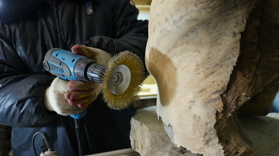 woodworker with a sander in his hands next to a piece of natural wood, using a metal round brush to treat the surface of a wooden material, a tool for rough sanding wood