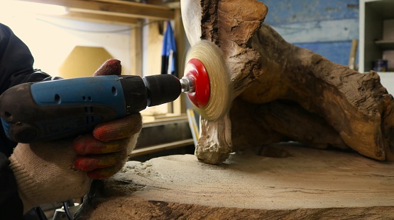 A grinding machine with iron bristles in the hands of a craftsman polishing a piece of whole wood material in the workshop, the woodworker cleans a metal brush on a drill of uneven natural wood