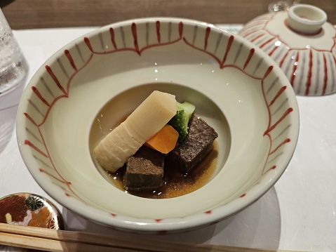 This is a photo of a traditional Japanese dish served in a white ceramic bowl with an elegant red pattern around the rim. The dish contains a clear broth with several ingredients including a piece of beef that appears to be braised or stewed, a slice of carrot, a piece of green vegetable which could be a type of leafy green or bell pepper, and a chunk of white vegetable that could be daikon radish. The dish is presented beautifully with the ingredients carefully placed in the center of the bowl. In the background, we can see a portion of a soy sauce pot and a pair of chopsticks indicating that this meal is likely part of a Japanese dining setting.