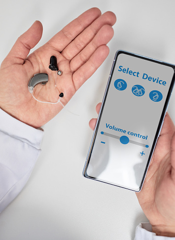 Doctor audiologist showing smartphone app for adjusting hearing aids holding smartphone in one hand and hearing aids in other. Manage hearing aid settings via smartphone