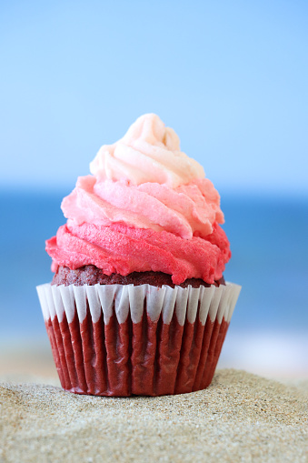 Stock photo showing close-up view of a single freshly baked, homemade, red velvet cupcake in paper cake case on a pile of sand, against a seaside with breaking waves background. The cup cake has been decorated with a swirl of ombre effect pink piped icing. Birthday holiday and vacation concept.