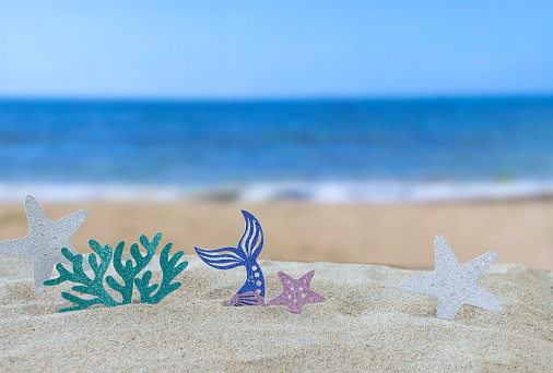 Stock photo showing close-up view of multi coloured seaside influenced cardboard cut out shapes on a pile of sand, against a seaside with breaking waves background. Holiday and vacation concept.