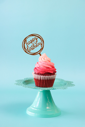 Stock photo showing close-up view of a single freshly baked, homemade, red velvet cupcake in paper cake case on cake stand. The cup cake has been decorated with a swirl of ombre effect pink piped icing.