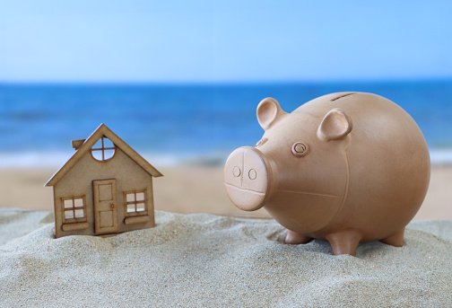 Stock photo showing close-up view of model coastal house besides piggy bank on sand pile on a sunny beach with the sea and clear blue sky in the background. Real estate, vacation home and beach hut rental concept.