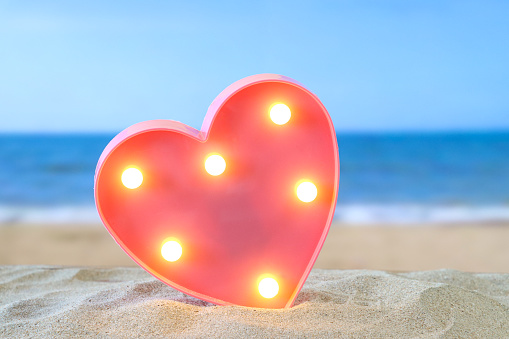 Stock photo showing close-up view of pink, heart shaped box lined with mounted illuminated, light bulbs on a pile of sand, against a seaside with breaking waves background. Romantic holiday and vacation concept.