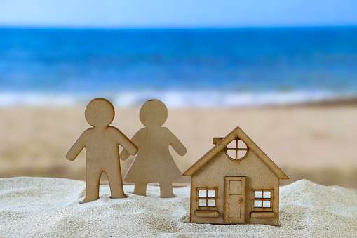 Stock photo showing close-up view of model wood cutout man and woman (heterosexual couple) near a wooden cutout, model coastal house on a sand pile on a sunny beach with the sea and clear blue sky in the background. Real estate, vacation home and beach hut rental concept.