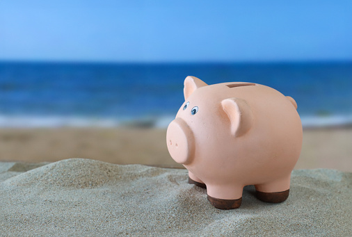 Stock photo showing close-up view of ceramic piggy bank on sand pile on a sunny beach with the sea and clear blue sky in the background. Holiday and vacation savings concept.