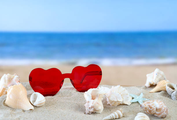 Close-up image of red, heart-shaped framed sunglasses on golden sand surrounded by heap of seashells and starfish, sunny sandy beach, waves breaking at low tide, sea and coastline, focus on foreground, copy space